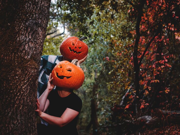 Zwei Personen verstecken sich hinter einem Baum, ihre Köpfe durch geschnitzte Kürbislaternen ersetzt, umgeben von herbstlichem Wald. | © AdobeStock/Cavan