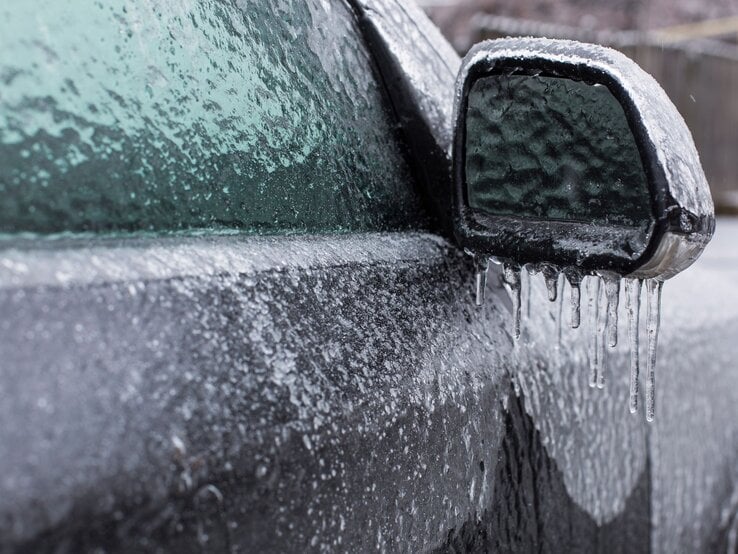 Außenspiegel und Fenster eines Autos komplett von einer dicken Eisschicht bedeckt, an der Eiszapfen hängen.