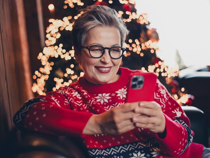 Eine grauhaarige Frau mittleren Alters mit Brille lächelt, während sie auf einem Sessel in rotem Weihnachtspullover ein Smartphone hält.