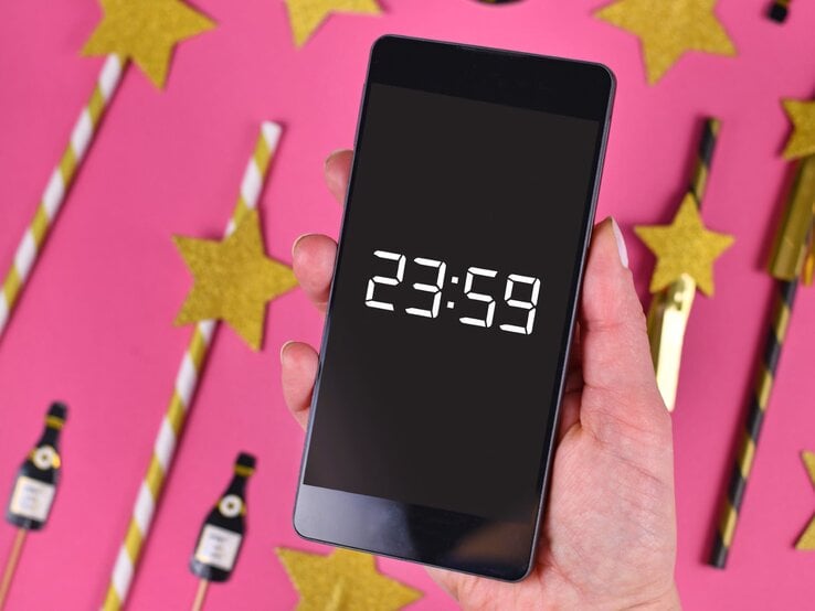 Silvestersprüche: Eine Hand mit rosa lackierten Nägeln hält ein Smartphone mit 23:59-Anzeige, umgeben von goldenen Sternen und Partydekorationen auf pinkem Hintergrund.