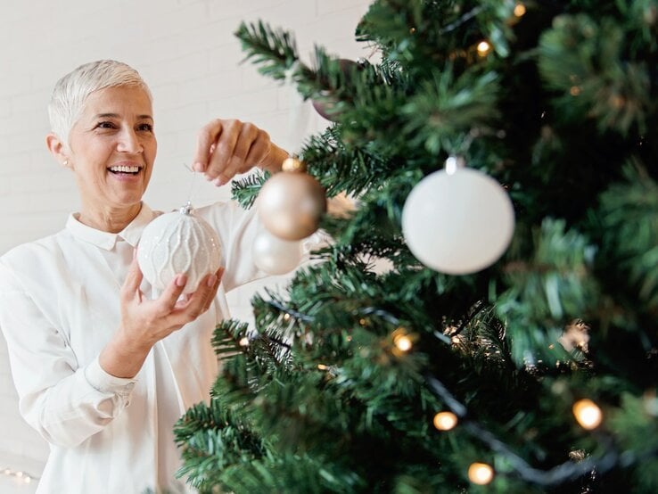 Eine lächelnde ältere Frau mit kurzen grauen Haaren hängt eine weiße Christbaumkugel an einen festlich geschmückten Tannenbaum.