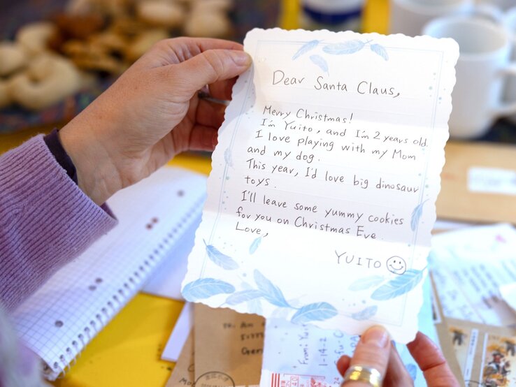 Eine Hand in einem fliederfarbenen Pullover hält einen handgeschriebenen Weihnachtsbrief an den Weihnachtsmann, im Hintergrund Kekse und Briefe. | © BILD der FRAU 