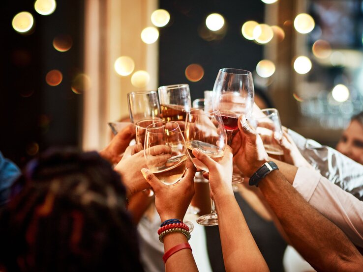 Mehrere Hände heben Gläser mit Wein und Sekt für einen fröhlichen Toast vor einem Hintergrund aus warmen Lichtern.