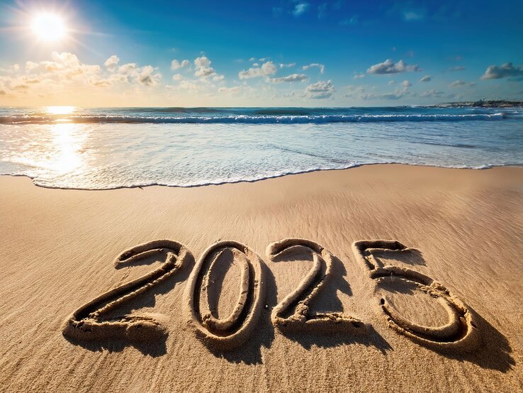 Die Zahl „2025“ ist in nassen Sand am Strand geschrieben, während Sonnenstrahlen das ruhige Meer im Hintergrund erhellen.