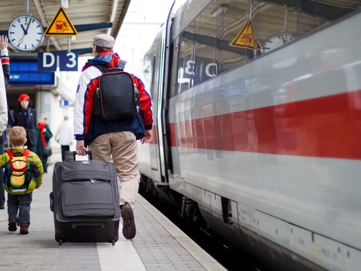 Ein Mann mit grauer Mütze und roter Jacke zieht einen Koffer, begleitet von einem Jungen mit gelbem Rucksack, auf einem Bahnsteig, an dem ein ICE der Deutschen Bahn hält.