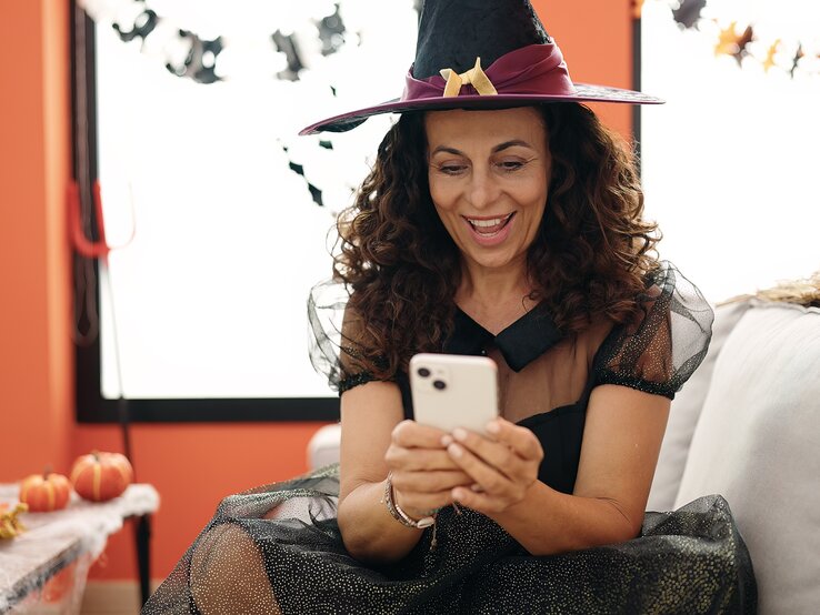 Halloween Sprüche Ein festlich dekorierter Raum mit Kürbissen, Kerzen und Spinnweben, dazu eine Person mit Hexenkostüm und Smartphone in der Hand.