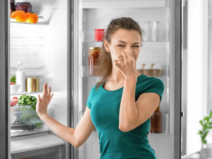 Junge Frau mit grünem Shirt hält sich die Nase zu und steht vor einem offenen, gefüllten Kühlschrank.