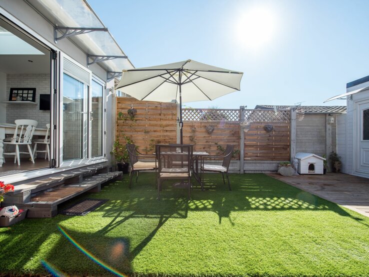 Sonnige Terrasse mit grüner Kunstrasenfläche, Tisch und Stühlen unter einem großen Sonnenschirm, daneben ein offenes Wohnzimmer.