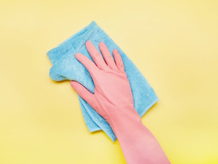 Eine Hand im rosa Gummihandschuh wischt mit einem blauen Microfaser Putzlappen über einen gelben Hintergrund.