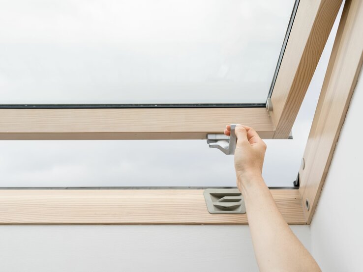Eine Hand öffnet ein Dachfenster aus Holz mit einem grauen Griff an einem bewölkten Tag.