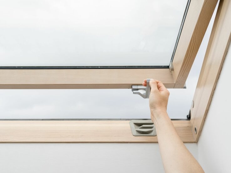 Eine Hand öffnet ein Dachfenster aus Holz mit einem grauen Griff an einem bewölkten Tag.