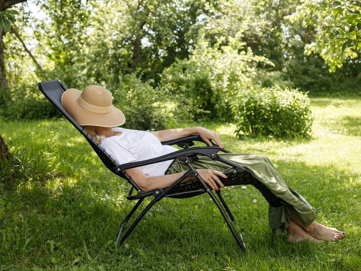 Barfüßige Frau mit Strohhut und grüner Hose ruht sich auf einem Liegestuhl unter einem Baum im sonnigen Garten aus.