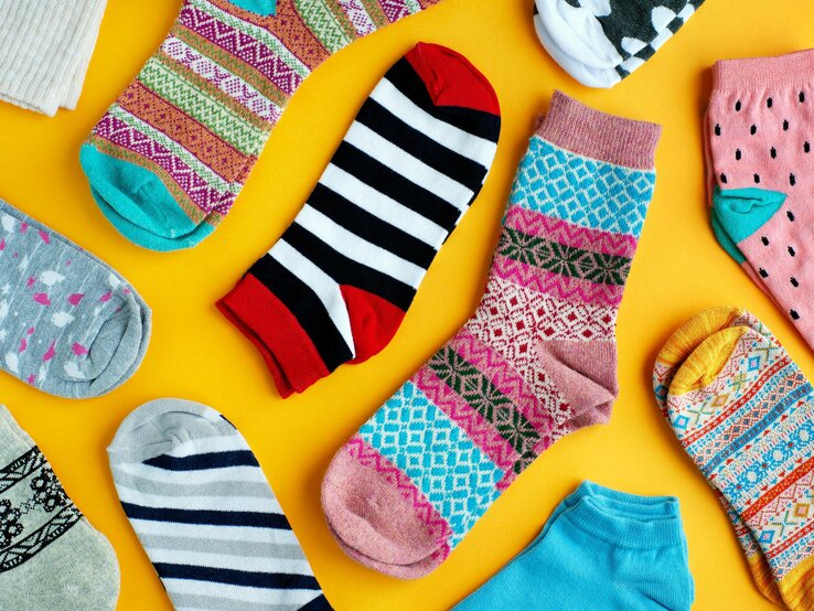 Bunte Auswahl verschiedener Socken auf einem leuchtend gelben Hintergrund. Die Socken weisen vielfältige Muster und Farben auf, darunter Streifen, Punkte, und geometrische Designs in Farben von Rot und Schwarz bis hin zu Pastelltönen.