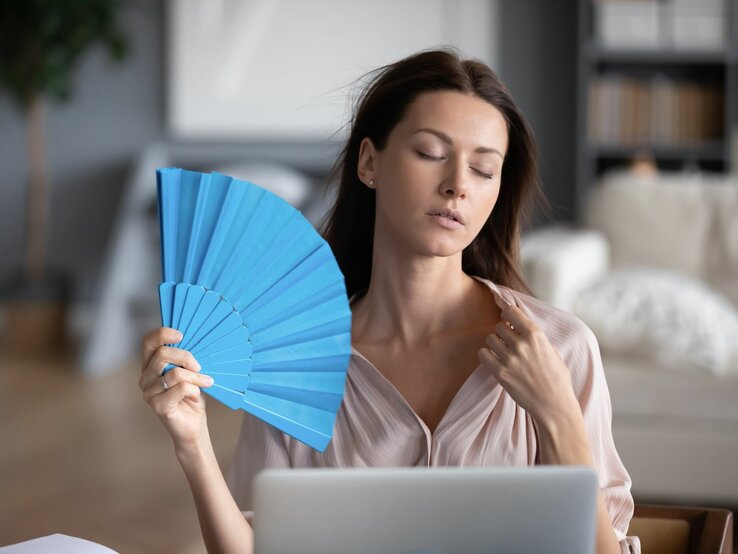 Das Bild zeigt eine Frau, die an einem Laptop sitzt und sich mit einem blauen Fächer Luft zufächelt. Sie wirkt erschöpft.