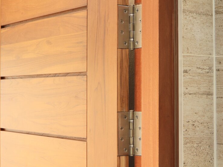 Holztür mit sichtbarem Metallscharnier, das an einer beige gekachelten Wand montiert ist.