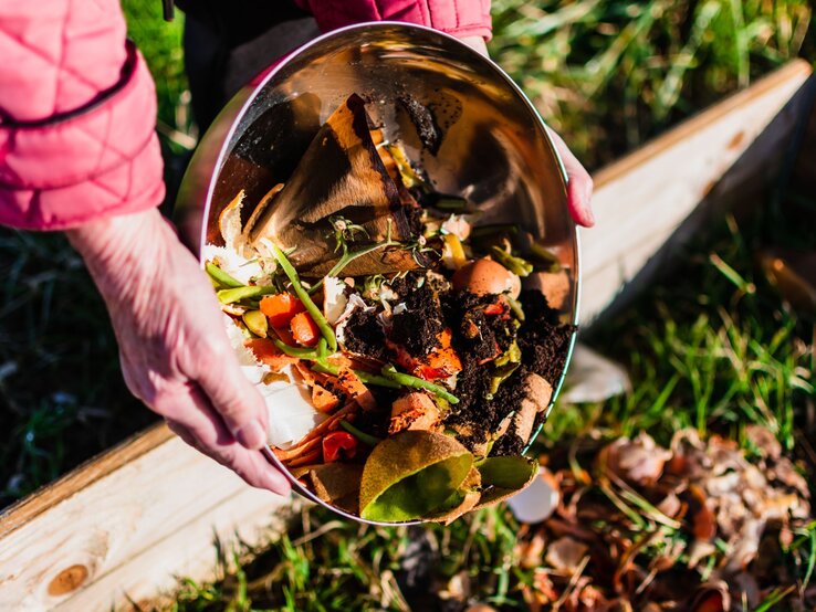 Eine Person in rosa Jacke entleert eine Schüssel mit Gemüseabfällen und Erde auf ein Beet im sonnigen Garten. | © Shutterstock/Reflexpixel