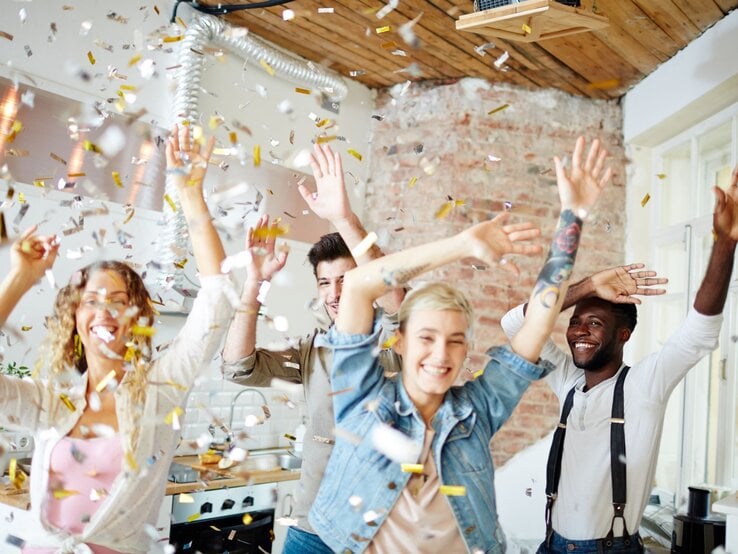 Vier junge Menschen feiern ausgelassen in einem loftartigen Raum mit Ziegelwänden eine Party, während Konfetti in der Luft schwebt.