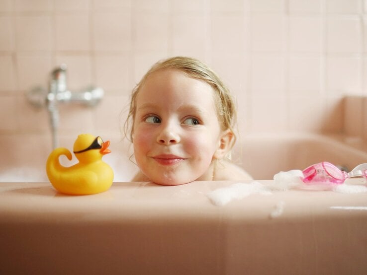 Ein blondes Kleinkind mit verschmitztem Lächeln schaut in einer rosafarbenen Badewanne neben einer gelben Gummiente hervor.