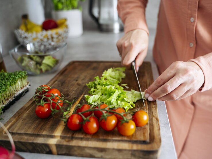 Eine Person in rosa Bluse schneidet auf einem Holzbrett frischen grünen Salat neben leuchtend roten Tomaten.