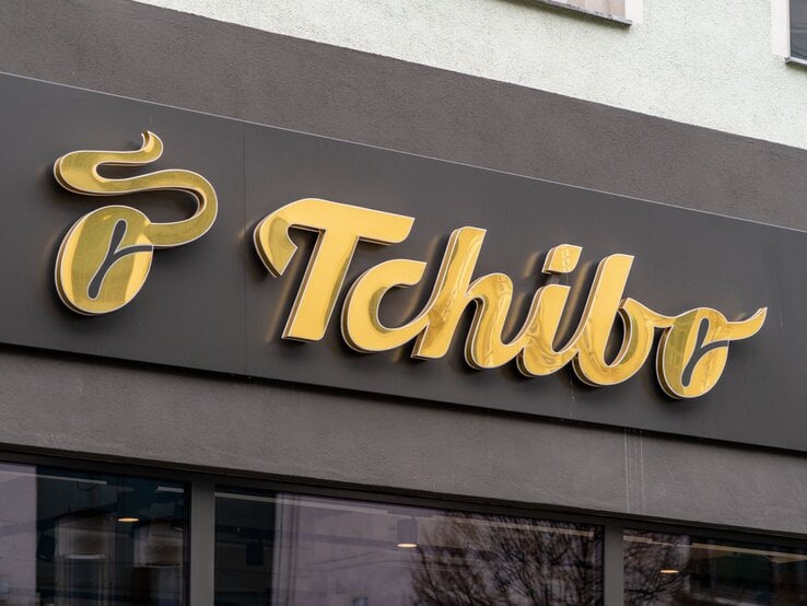 Goldenes Tchibo-Logo auf schwarzem Hintergrund an einer Hausfassade, unterhalb Fenster mit Spiegelung von Bäumen.