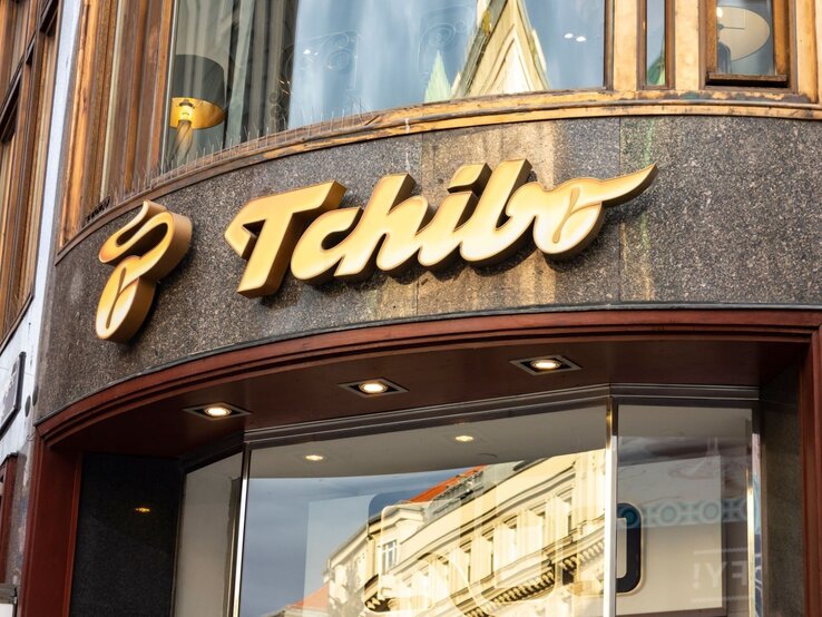 Eingang einer Tchibo-Filiale mit großem goldenen Logo auf dunkler Fassade und spiegelnder Glasscheibe.