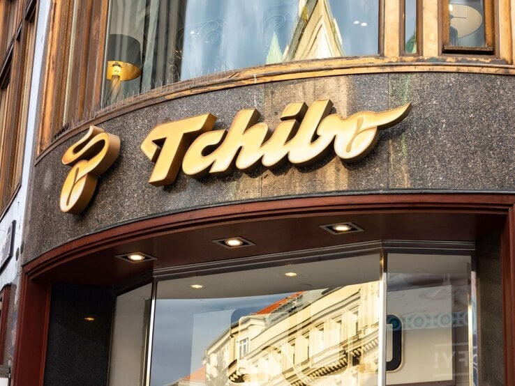 Eingang einer Tchibo-Filiale mit großem goldenen Logo auf dunkler Fassade und spiegelnder Glasscheibe.