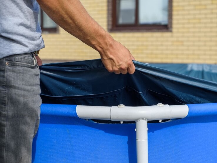 Ein Mann in grauem T-Shirt und Jeans spannt eine blaue Poolfolie über einen Aufstellpool vor einem gelben Backsteingebäude.