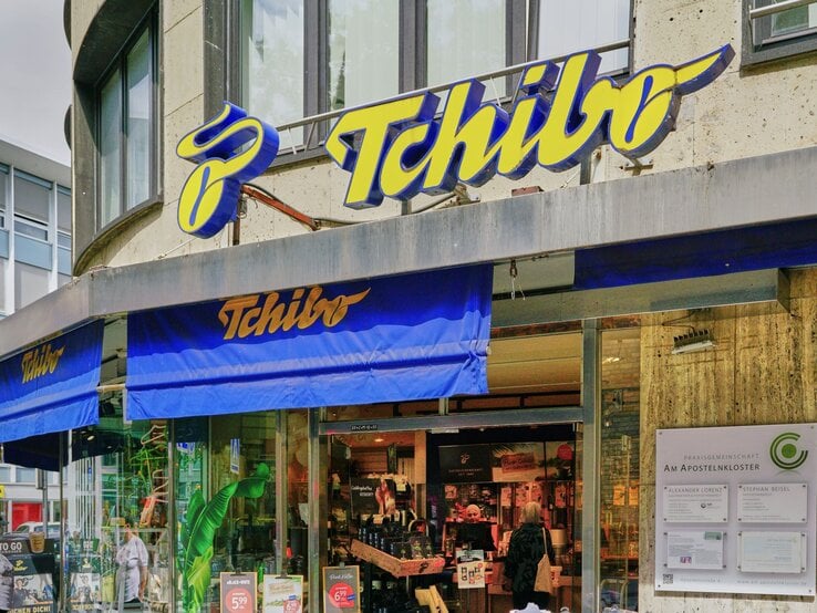 Eingangsbereich einer Tchibo-Filiale mit blauen Markisen und großem gelbem Logo, Passantin mit Tasche tritt gerade in den Laden ein.