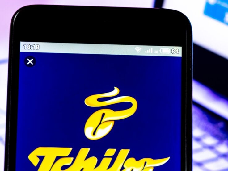 Tchibo-Logo auf einem Smartphone-Bildschirm vor einem unscharfen Laptop, der einen Kursverlauf anzeigt.