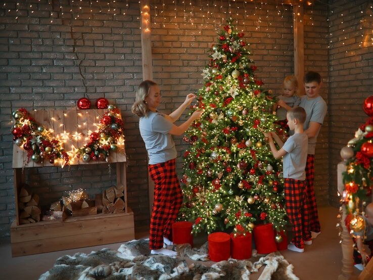 Eine lächelnde Familie schmückt in karierten Schlafanzügen einen leuchtenden Weihnachtsbaum vor einer Backsteinwand mit Lichterketten.