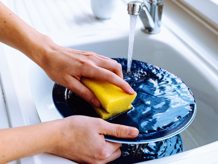 Eine Person spült in einem weißen Waschbecken mit einem gelben Schwamm einen nassen, blauen Teller unter fließendem Wasser ab. | © Shutterstock/larisa Stefanjuk