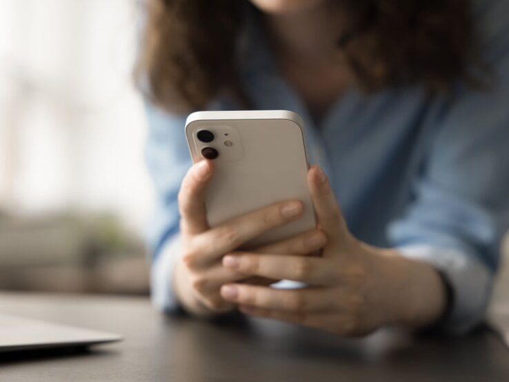 Eine Frau in blauer Bluse hält ein weißes Smartphone in beiden Händen und blickt konzentriert darauf. | © Shutterstock/fizkes