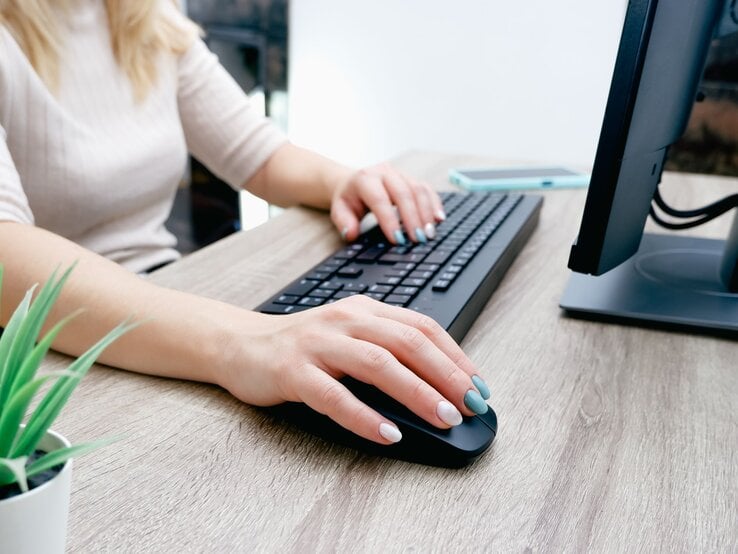 Eine Frau mit pastellfarben lackierten Nägeln bedient an einem Holztisch eine Computermaus und eine schwarze Tastatur. | © Shutterstock/Fire-n