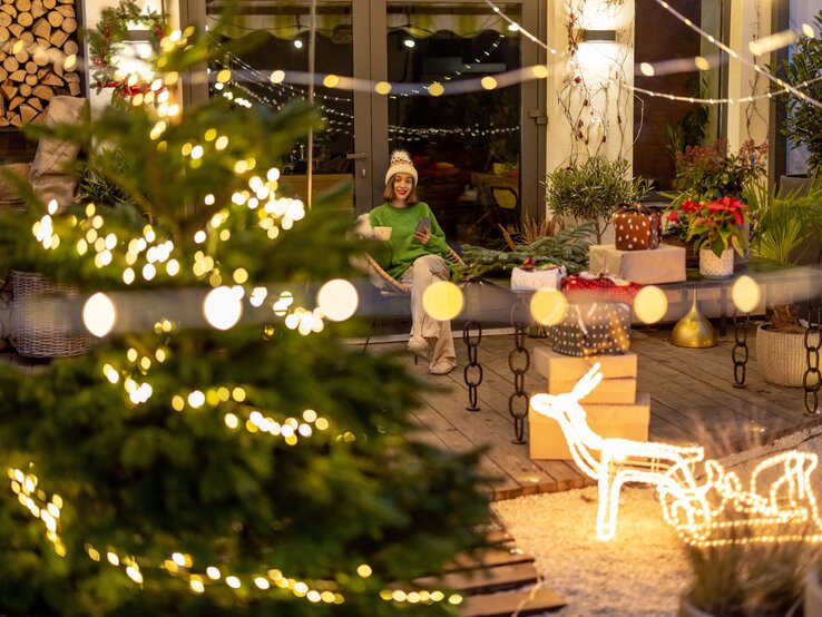 Eine junge Frau mit weißer Mütze und grünem Pullover sitzt lächelnd auf einer Terrasse, umgeben von Weihnachtsdeko und Geschenken.