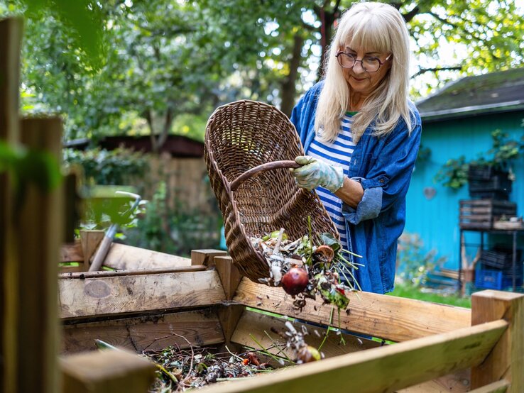 Eine ältere Frau mit blondem Haar und Brille leert lächelnd einen Korb mit Gartenabfällen in einen Kompostbehälter in grünem Garten.