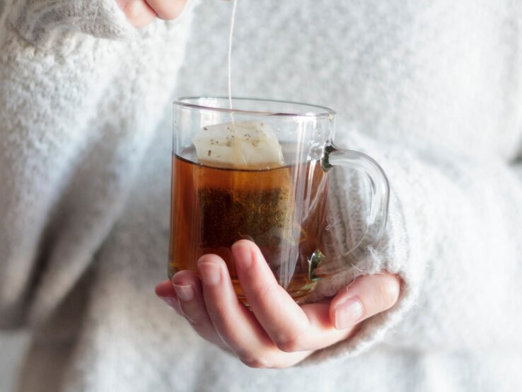 Eine Hand in einem flauschigen weißen Pullover hält eine Tasse heißen Tee, während ein Teebeutel langsam zieht.