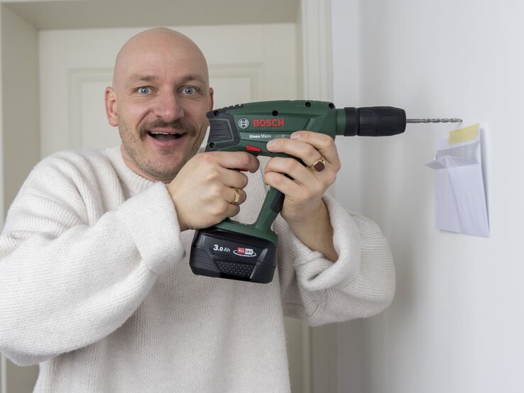 Ein fröhlicher Mann mit blauem Blick und Schnurrbart bohrt mit einer Bosch-Bohrmaschine nahe eines angeklebten Umschlags in eine Wand.