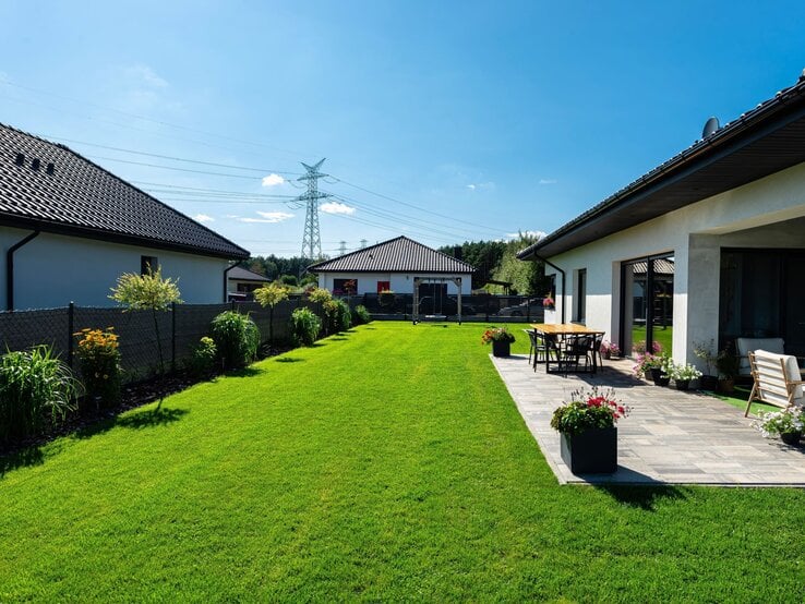 Sonniger Garten mit gepflegtem Rasen, moderner Terrasse mit Holztisch und Stühlen, umgeben von Blumen und zwei weißen Einfamilienhäusern. | © Shutterstock/Kinek00