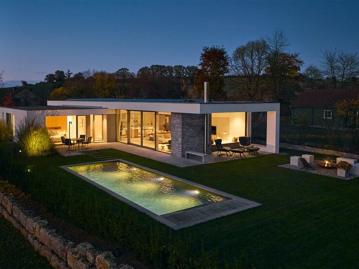 Stilvolles Flachdachhaus mit gemütlich erleuchteten Innenräumen, Pool und Sitzbereich im weitläufigen Garten am Abend.