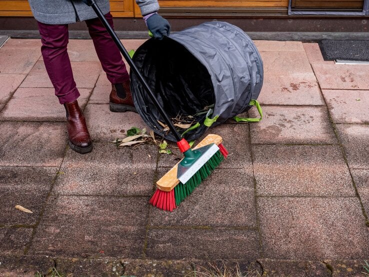 Eine Person in bordeauxroter Hose und braunen Stiefeletten fegt Laub mit einem Besen auf eine umgekippte Gartentasche auf der Terrasse.