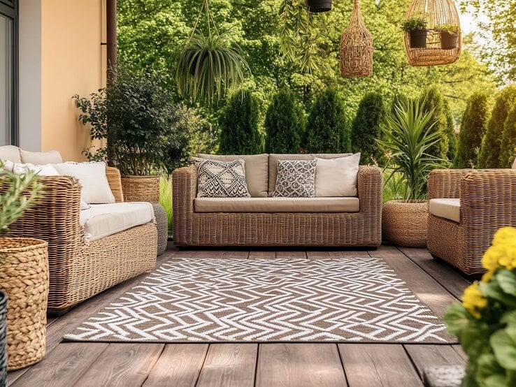 Stilvolle Outdoor-Lounge mit Rattansofas, Holzboden und Outdoor- Teppich, eingerahmt von üppigem Grün und dekorativen Hängepflanzen.