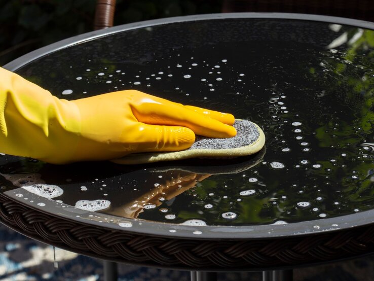 Eine Hand mit gelbem Gummihandschuh reinigt mit einem Schwamm einen nassen Glastisch, auf dem Seifenblasen schimmern.