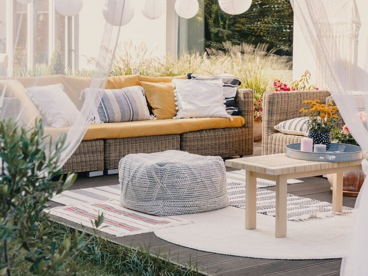 Gemütliche Terrasse mit Rattansofa, Holztisch und gemusterten Outdoorteppichen in Beige, Weiß und Rot, umgeben von Pflanzen