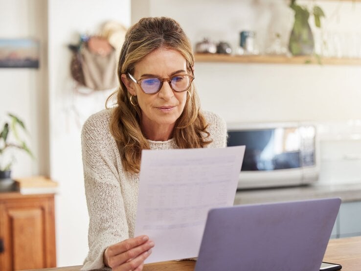 Frau, die konzentriert ein Dokument liest, während sie an einem Laptop sitzt. Sie trägt eine Brille und ist in einer gemütlichen Wohnküche. Im Hintergrund sind Mikrowelle und Pflanzen zu sehen.