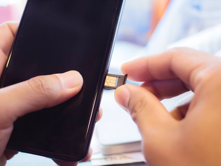 Eine Hand setzt eine goldene SIM-Karte in den geöffneten Kartenslot eines schwarzen Smartphones ein, im Hintergrund ein heller Schreibtisch.