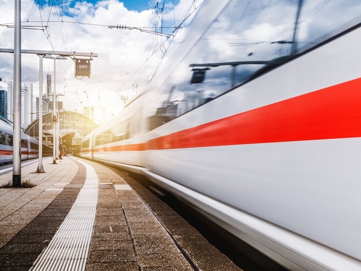 Ein ICE-Zug rauscht mit hoher Geschwindigkeit an einem Bahnsteig vorbei, während die Sonne hinter einer modernen Bahnhofshalle aufgeht.