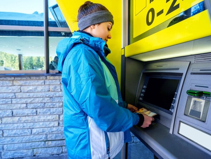 Eine junge Frau in blauer Winterjacke und Stirnband hebt an einem Geldautomaten Geld ab, während sich ihr Gesicht konzentriert nach unten neigt.