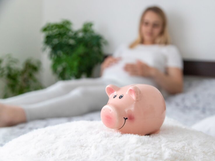 Ein rosa Sparschwein mit lachendem Gesicht steht auf einer flauschigen weißen Decke, im Hintergrund liegt eine schwangere Frau in Weiß.