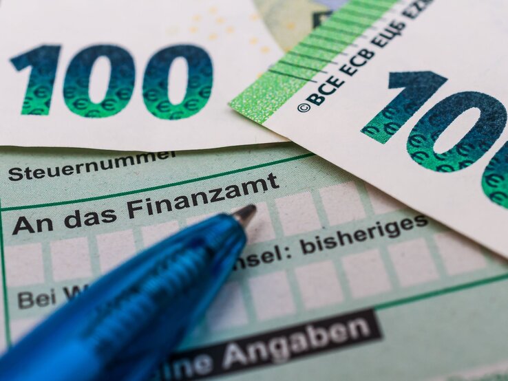 Zwei 100-Euro-Scheine liegen auf einem Steuerformular mit der Aufschrift "An das Finanzamt", daneben ein blauer Kugelschreiber.