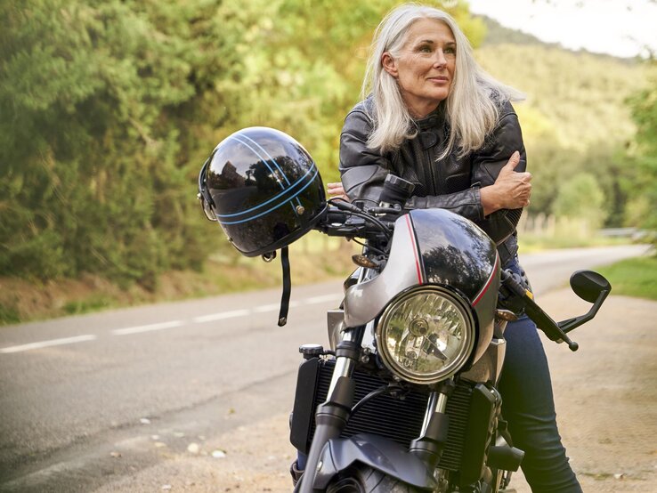 Zu sehen ist eine Frau mittleren Alters mit weißen Haaren, die sich lässig über ihr Motorrad lehnt